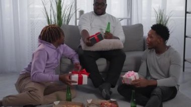 Holiday present. Friendly meeting. Happy black men. Relaxed male friends holding gift boxes talking together with beer and pizza sitting light room interior.