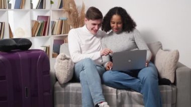 Choosing tour. Happy couple. Online order. Positive beloved man and woman looking laptop for journey with packed suitcase in light room interior.