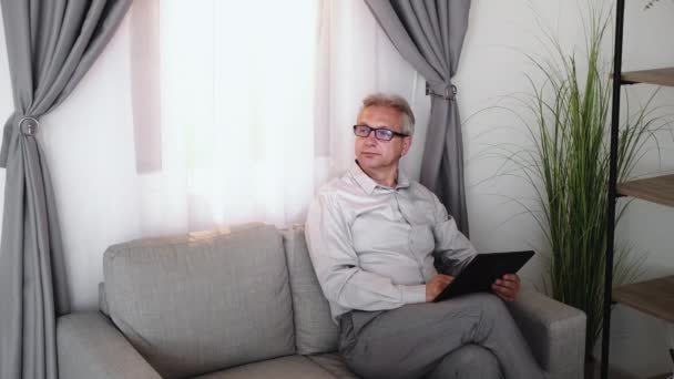 数字休闲 现代技术 在线业余爱好 头戴眼镜的聪明而沉思的中年男子 在家里光线明亮的客厅里 用平板电脑在沙发上做着创造性的工作 — 图库视频影像