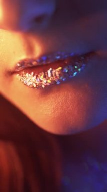 Vertical video. Glitter lips. Neon light girl. Glamour beauty. Cropped closeup of red blue color glow woman face with artistic makeup shimmering metallic skin on dark.