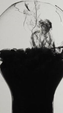 Vertical video. Black smoke. Ink shot. Dark energy. Toxic fume cloud filling broken light bulb glass lamp on white abstract background.