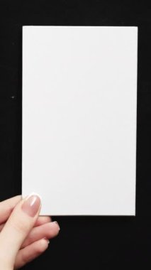 Vertical video. Blank card. Information letter. Female hands showing white mockup paper sheet with empty space on black background set of 2 loop.