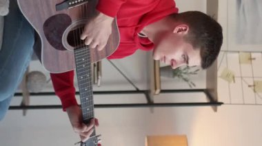 Vertical video. Male guitarist. Musical lesson. Home play. Talented dreamy man playing chords on acoustic guitar sitting couch light room interior.