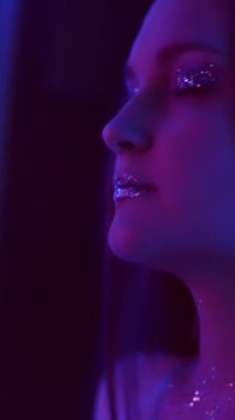 Vertical video. Neon girl. Glitter makeup. Nightclub beauty. Purple color light relaxed woman with sparkling face skin eyeshadow lips dancing with closed eyes on dark.