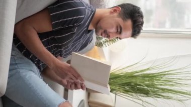 Vertical video. Home leisure. Reading man. Enjoying weekend. Happy casual guy sitting comfortable couch with interesting book in light room interior.