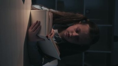 Vertical video. Night fairy tale. Kid evening leisure. Scared overwhelmed little girl reading book spooky story at desk late at dark home interior.