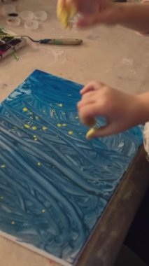 Vertical video. Kid painting. Art class. Bonding leisure. Mother little child hand fingers sprinkling blue color abstract artwork with yellow paint.