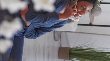 Vertical video. Mobile communication. Happy woman. Home leisure. Cheerful female enjoying talk by smartphone sitting floor light room interior.