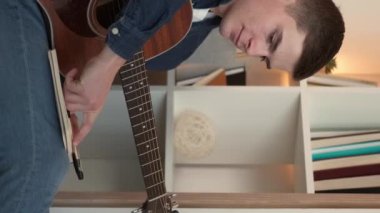 Vertical video. Song writing. Male composer. Home leisure. Creative man making notes in journal playing guitar sitting light room interior.