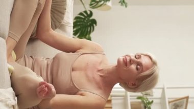 Vertical video. Peaceful meditation. Inspired woman. Healing energy. Smiling relaxed middle-aged lady sitting yoga pose on sofa in light home interior.