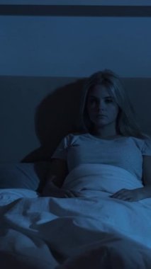 Uyku sorunu. Gece uykusuzluğu. Yorgunluk depresyonu. Üzgün, yorgun, dalgın bir kadın karanlık bir yatak odasında tek başına uyanık oturuyor. Dikey video.
