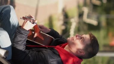 Vertical video. Street guitarist. Male musician. Artistic life. Talented man playing acoustic guitar sitting bench in sunny park.