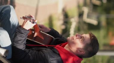Vertical video. Street guitarist. Male musician. Artistic life. Talented man playing acoustic guitar sitting bench in sunny park.