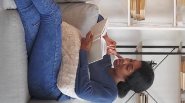 Vertical video. Book leisure. Calm woman. Home rest. Happy lady enjoying reading sitting couch light room interior.