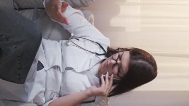 Vertical video. Diagnostic radiology. Injury diagnosis. Medical healthcare. Disturbed woman doctor examining x-ray film consulting patient on phone call at hospital.