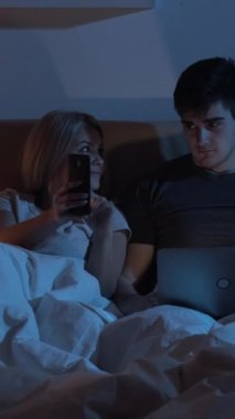 Gece online. Gadget çifti. Ev dijital eğlencesi. Hevesli kadın yorgun adam karanlık yatak odasında cep telefonu laptopu kullanıyor. Dikey video.