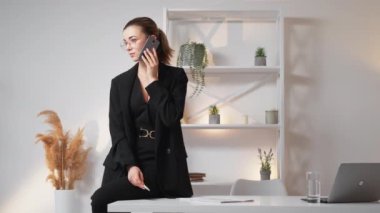 Mobile communication. Elegant woman. Office routine. Positive business lady talking smartphone sitting desk in light room interior.