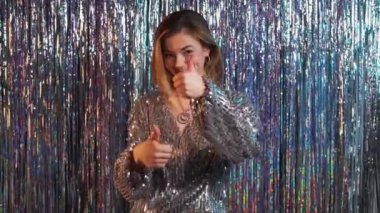 Party joy. Happy woman. Great celebration. Festive smiling lady showing thumbs up dancing in light blue silver cascade curtain background.