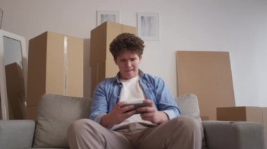 Game addiction. Nervous man. Home moving. Stressed angry guy loosing playing smartphone sitting sofa and packed cardboard boxes in light room interior.