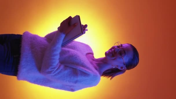 垂直ビデオ インスピレーションを得た計画 ネオンライトの女 幸せな夢のような女性作るノートパッドでメモを取る紫色のオレンジの背景 — ストック動画