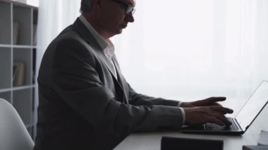 Business online. Digital marketing. Virtual communication. Confident smart middle-aged executive man working typing on laptop at light home office workplace with free space.