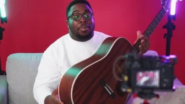 Music influencer video blog. Instrument presentation. Professional musician man recording acoustic guitar review speaking on camera sitting on couch in red studio.