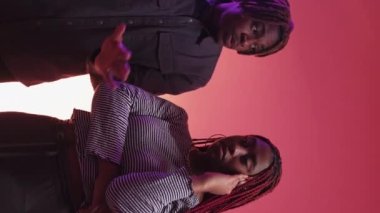 Vertical video. Boring date. Neon light couple. Inspired black man describing life to skeptic displeased woman feeling tired on purple peach pink background.