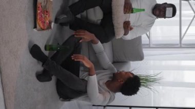 Vertical video. Home fun. Happy male friends. Enjoying meeting. Cheerful black men talking playing game guess who with pizza and beer sitting light room interior.