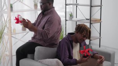 Pleasant gift. Curious black men. Holiday surprise. Positive inspired male friends holding shaking present boxes trying to guess what inside sitting light room interior.