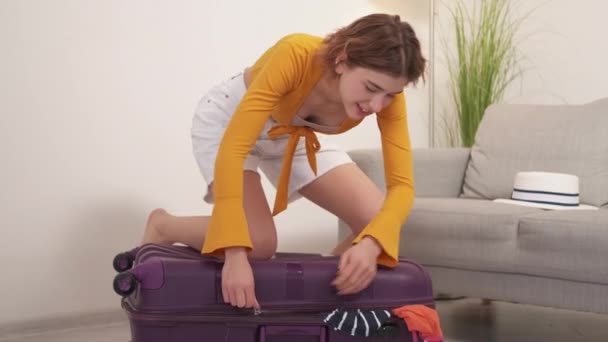 大きな荷物だ ストレスを受けた女性 旅行の準備だ かなり疲れている女性をパックしようとして服に滞在上のスーツケースジッパーそれで軽い部屋のインテリア — ストック動画