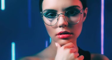 Optic fashion. 90s face. Retro look. Blue purple color neon light closeup portrait of confident teen girl model in glasses with artistic makeup on dark.