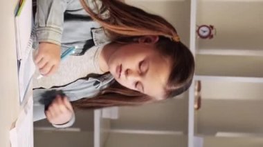 Vertical video. Kid homework. Primary school knowledge. Home learning. Diligent busy smart small girl studying writing exercise working at desk.