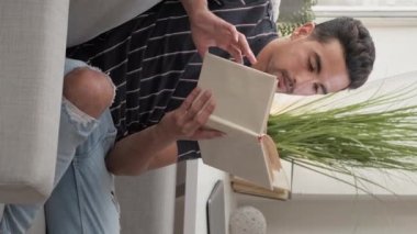 Vertical video. Enjoying reading. Curious man. Home rest. Handsome smiling guy sitting comfortable couch with interesting book in light room interior.