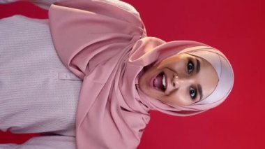 Vertical video. Wow surprise. Winner yes. Victory celebration. Portrait of impressed astonished happy smiling woman in hijab enjoying triumph on red.