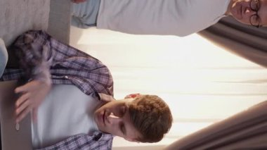 Vertical video. Private chat. Personal content. Disturbed excited shy teenager son hiding screen from dad closing laptop on couch at home living room.