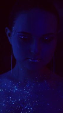 Vertical video. Neon portrait. Glitter skin. Night party look. Blue purple color light woman face with artistic makeup shimmering eyeshadow lips in mirror on dark.