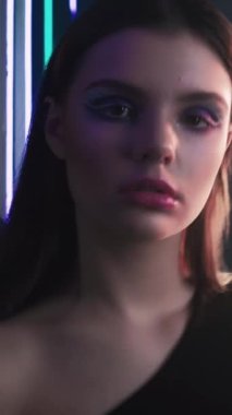 Vertical video. 90s model. Retro fashion. Old 2000s look. Portrait of confident teen girl face with artistic makeup posing in purple blue color neon light on dark black.