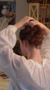 Vertical video. Artist lifestyle. Art therapy. Creative hobby. Inspired woman fixing hair with paintbrushes painting abstract picture on canvas at home studio.