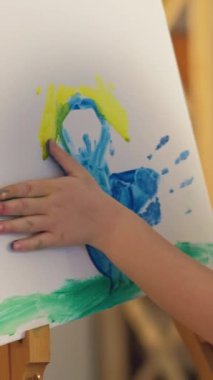 Vertical video. Kid art. Hand painting. Creative imagination. Unrecognizable inspired small girl fingers creating blue yellow color abstract artwork on easel.