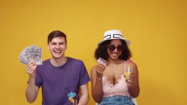 All-inclusive trip. Rich couple. Vacation budget. Happy man and woman showing fan of cash dollars and credit card drinking cocktails posing yellow background.