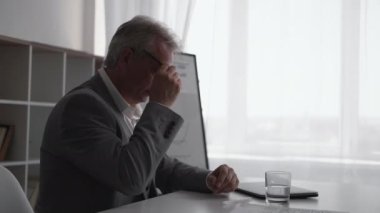 Work break. Office rest. Accomplished task. Tired relaxed satisfied senior business man taking off glasses drinking glass of water at light workplace interior with free space.