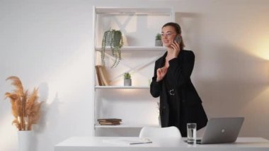 Distance communication. Talking woman. Work break. Positive business lady discussing interesting news by smartphone going in light office room interior.