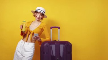 Mobile communication. Travelling woman. Departure waiting. Confident happy lady scrolling smartphone drinking cocktail leaning suitcase on yellow background.