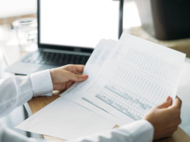 Accountant manager. Office work. Digital technology. Unrecognizable woman checking documents near laptop with blank screen in light room interior.