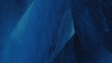 Dust mist. Glitter texture. Paint drip. Defocused blue color sparkling fluid ink smoke over cube angle in water on dark abstract background.
