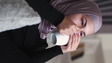 Vertical video. Coffee rest. Cozy weekend. Home leisure. Happy pensive relaxed woman in hijab with mug enjoying drinking hot beverage daydreaming on couch in living room.