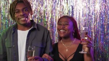 Christmas party. Greeting couple. Happy celebration. Joyful black man and woman saying toast with champagne on festive cascade curtain background.