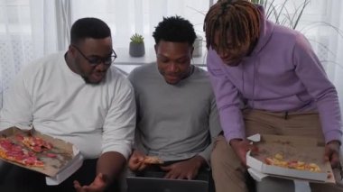 Online game. Relaxed male friends. Fast food dinner. Positive black men playing laptop discussing competition eating pizza in light room interior.