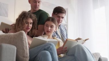 Book lover. Curious friends. Home club. Happy men and women discussing together interesting literature sitting sofa in light room interior.