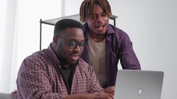 コンピュータ マスター 刺激を受けた男 解決策を 幸せな黒の男性の友人が見て満足作業ノートパソコンの座ってデスクライトルームインテリア — ストック動画